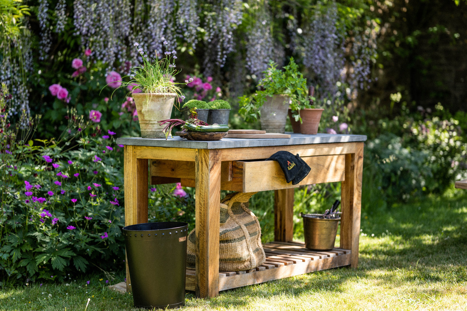Kadai outdoor kitchen table in a garden with potted herbs, vegetables, and a black kadai firepit accessory, surrounded by greenery and flowers.