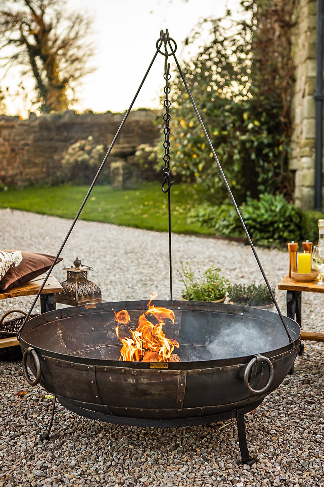 Outdoor tripod fire pit with a hanging chain above a lit fire, placed on a gravel patio surrounded by garden seating