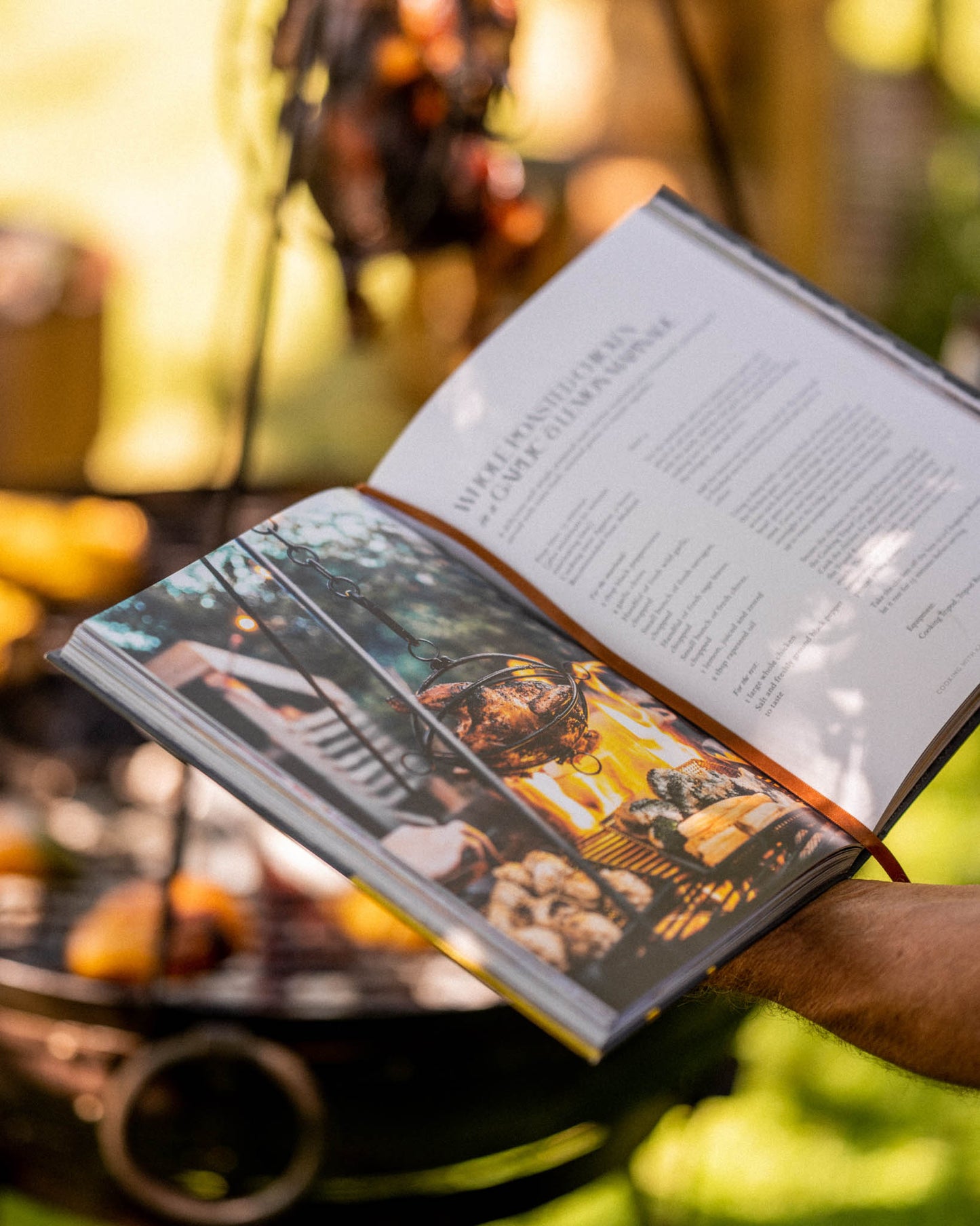 Open Kadai live fire cookbook held outdoors with recipe pages and fire pit cooking scene in background