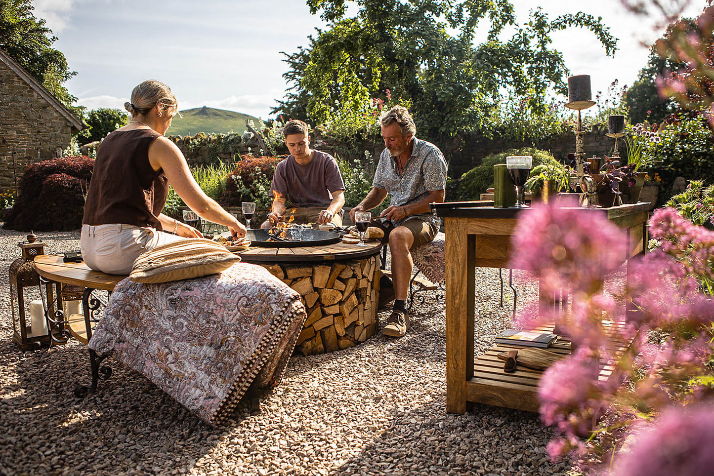 People sitting around a fire pit in a garden setting with flowers in the foreground.