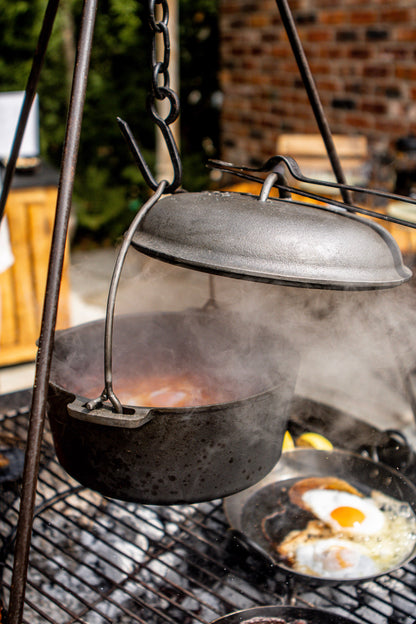 A dark cast iron Dutch oven hanging over a grill, with steam rising from the pot and a lid resting on top. A frying pan with two sunny-side-up eggs sits underneath on the grill, with an outdoor brick wall and greenery in the background.