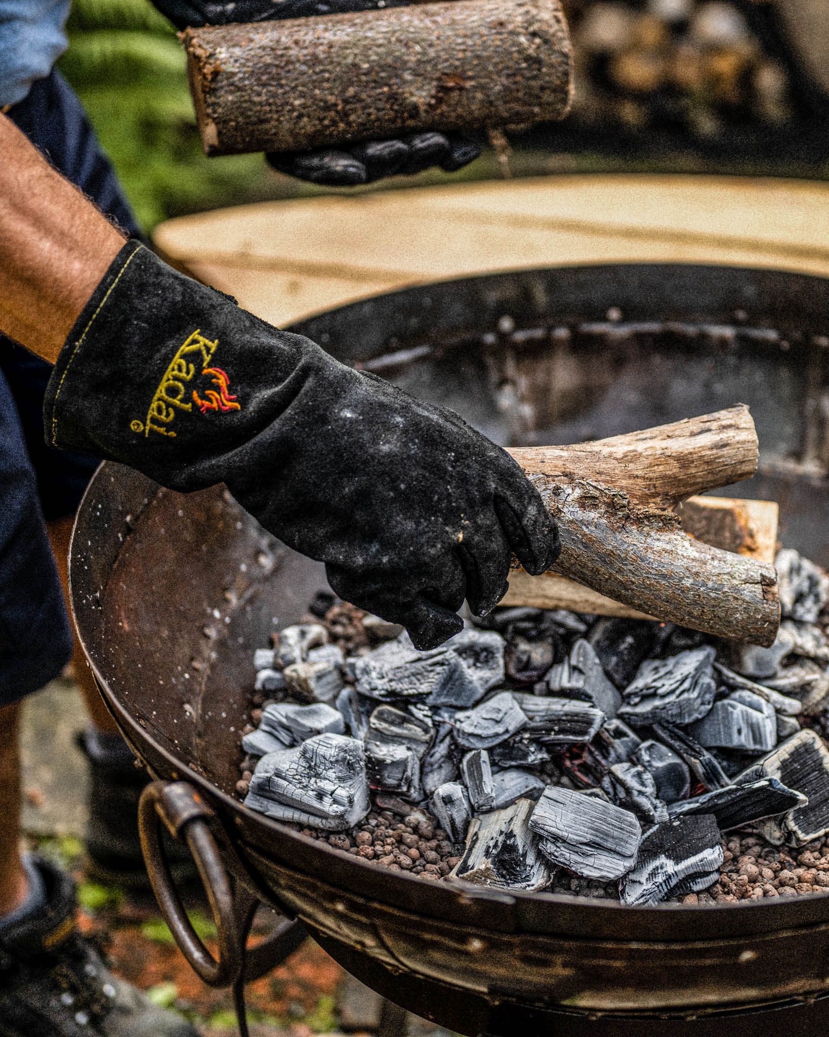 Person wearing heat-resistant kadai gloves placing firewood onto hot coals in a fire pit