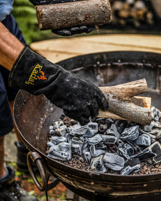 Person wearing heat-resistant kadai gloves placing firewood onto hot coals in a fire pit