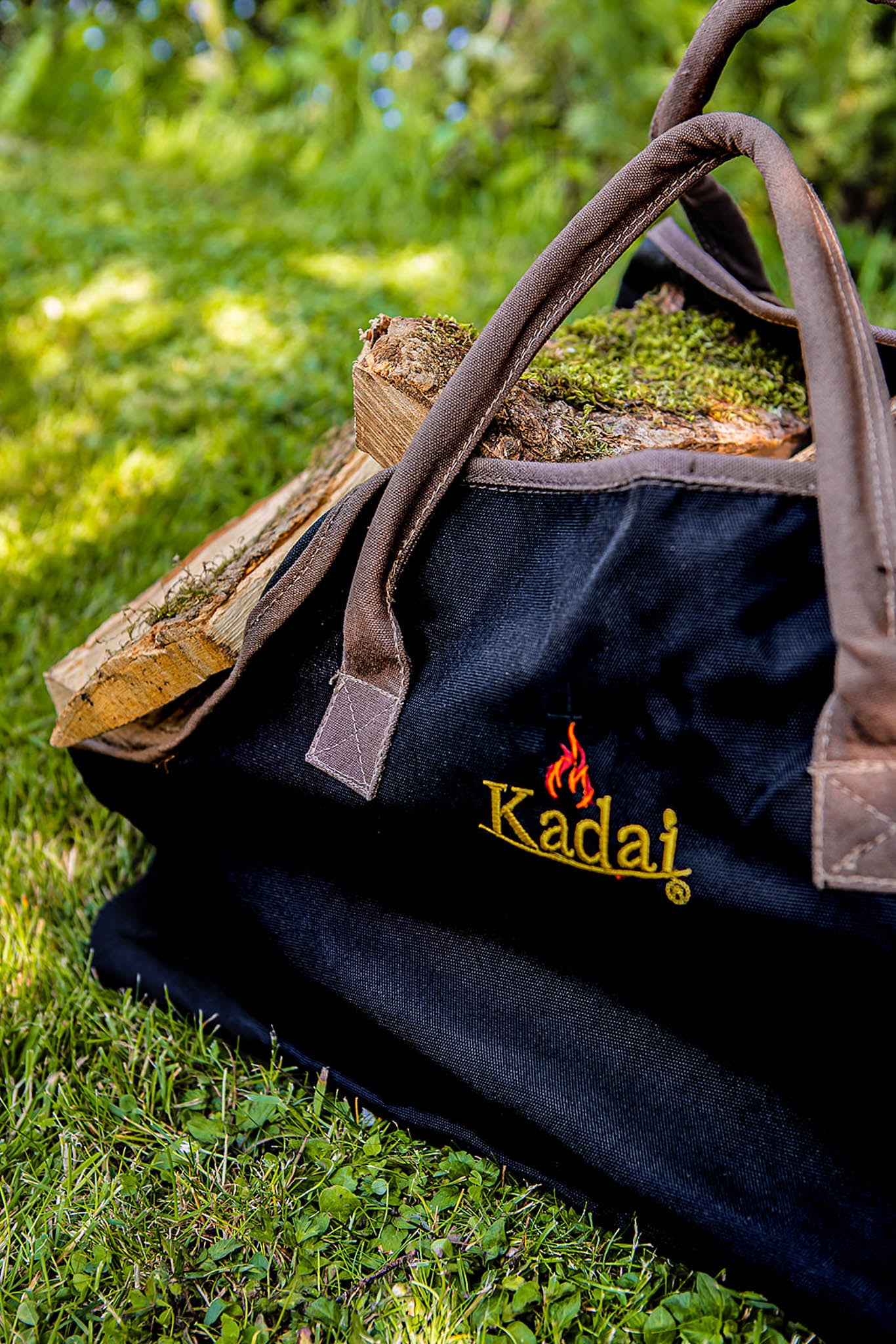 Black canvas log carrier filled with split firewood resting on grass in a garden setting.
