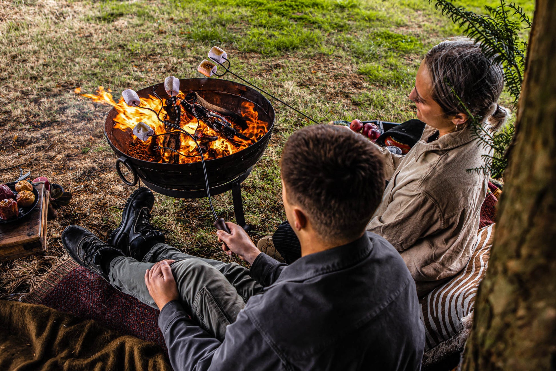 Outdoor campfire scene with friends toasting marshmallows above open flames using extended roasting forks on a Kadai fire bowl