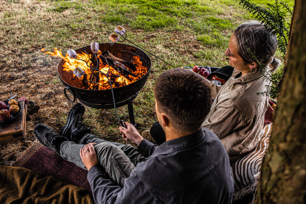 Outdoor campfire scene with friends toasting marshmallows above open flames using extended roasting forks on a Kadai fire bowl