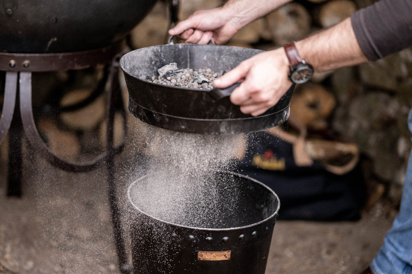 Close-up of a metal Kadai riddle shaking fine ash through a sieve while retaining charcoal and ceramic beads for reuse