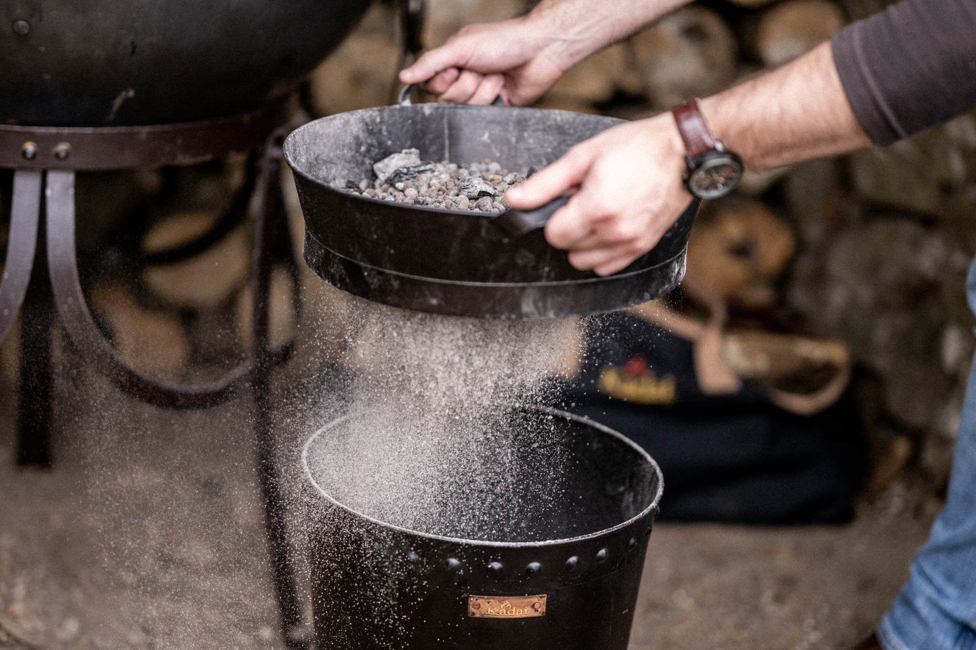 Close-up of a metal Kadai riddle shaking fine ash through a sieve while retaining charcoal and ceramic beads for reuse
