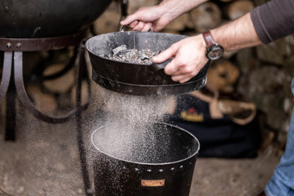 Close-up of a metal Kadai riddle shaking fine ash through a sieve while retaining charcoal and ceramic beads for reuse