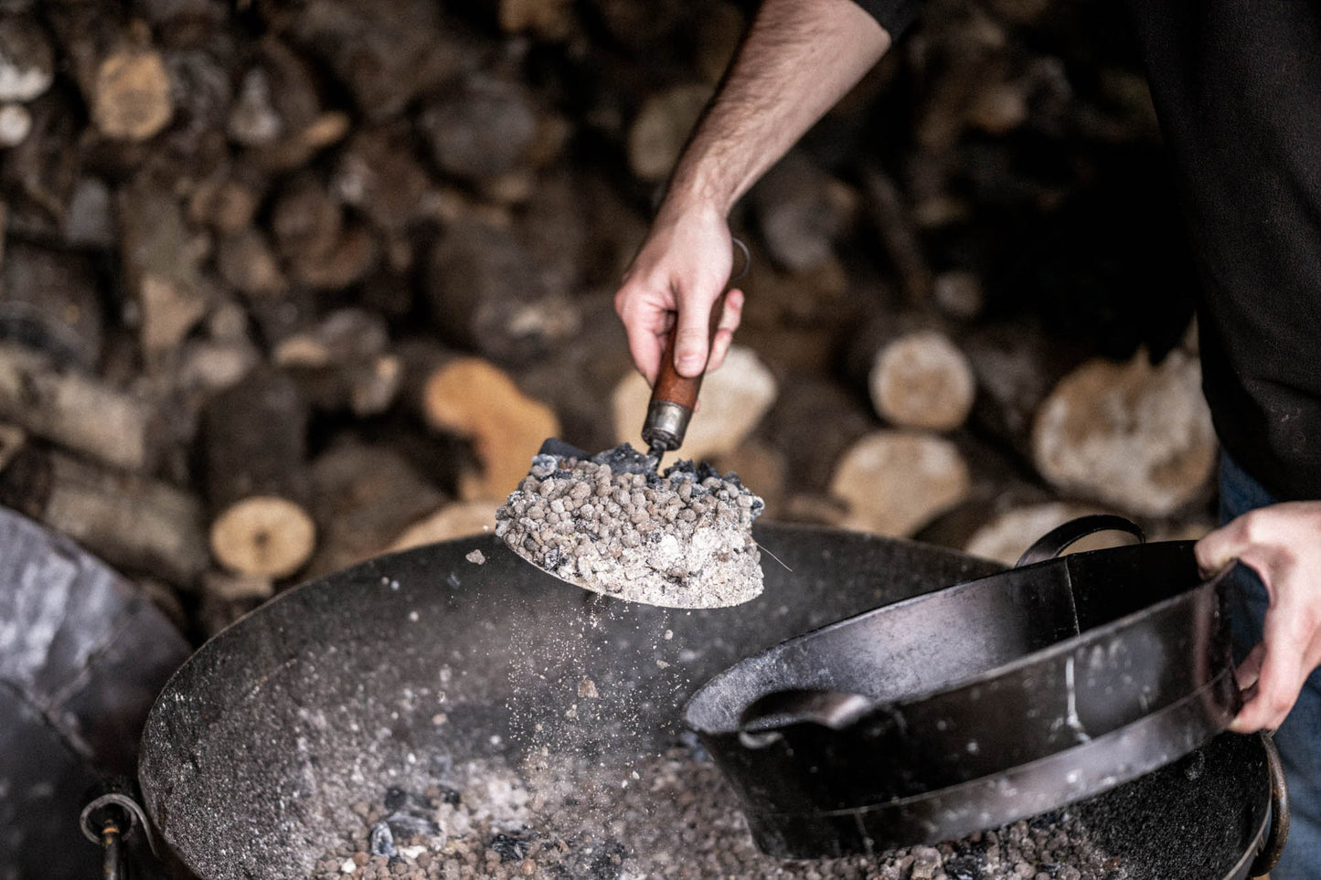 Person using a black metal Kadai shovel with a wooden handle to scoop ash and embers from a fire bowl into a metal ash pan.