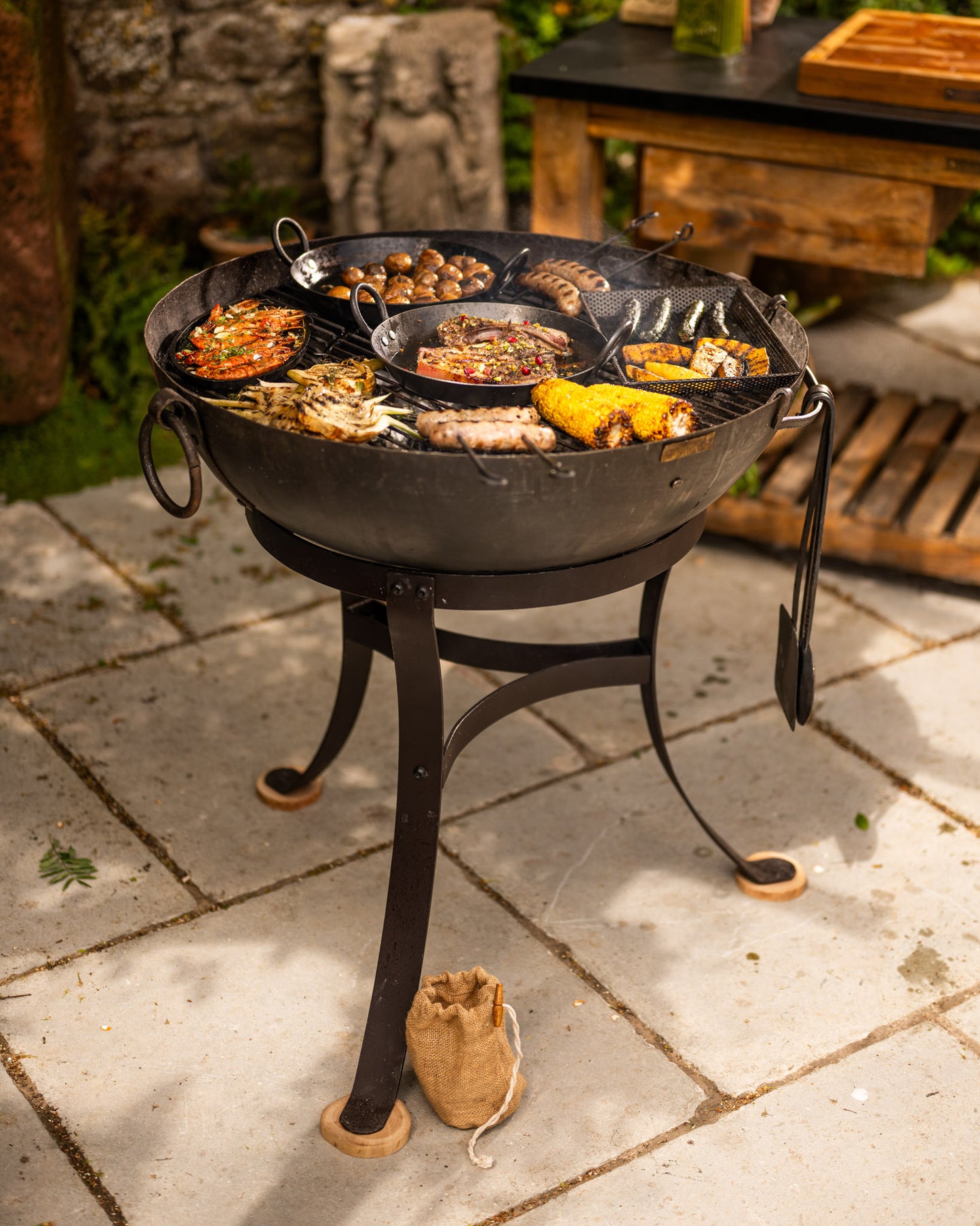Kadai fire pit with wooden feet protectors, featuring live-fire cooking, handcrafted barbecue accessories and outdoor entertaining on a stone garden patio