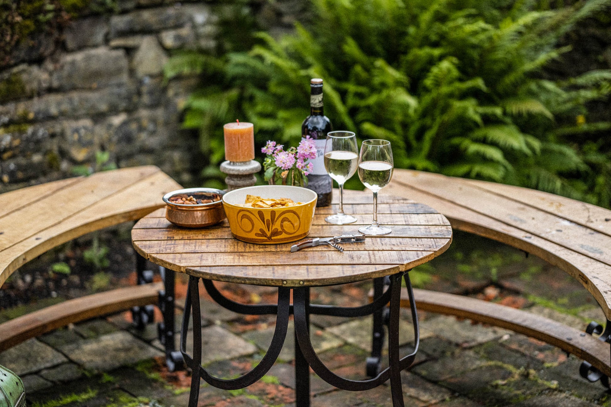 Natural wood patio table with drinks and food in a garden setting, framed by semi-circular seating