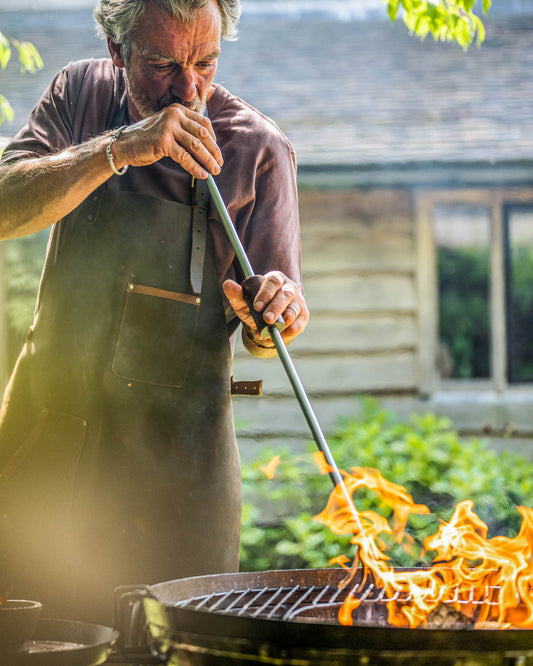 Man using a long metal blow poker to stoke flames in an outdoor fire pit while wearing a leather apron.