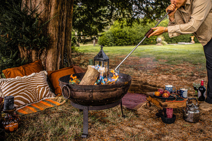 Person using a long metal blow poker with wooden handles to stoke flames in a Kadai fire bowl beside a cosy outdoor seating area with cushions, lanterns and drinks.
