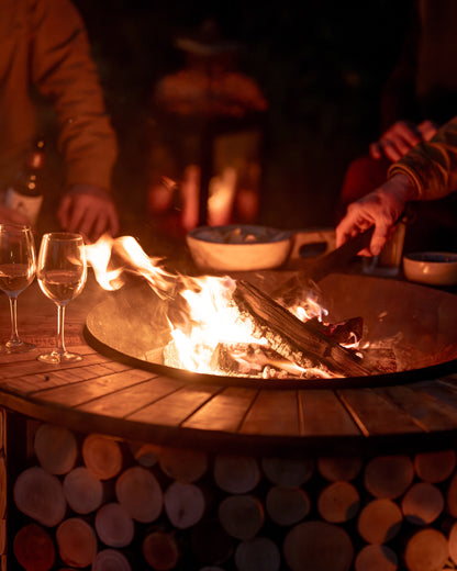 Round Kadai firebowl table with a blazing wood fire at the center, surrounded by people enjoying drinks and tending the flames during an evening outdoor gathering.