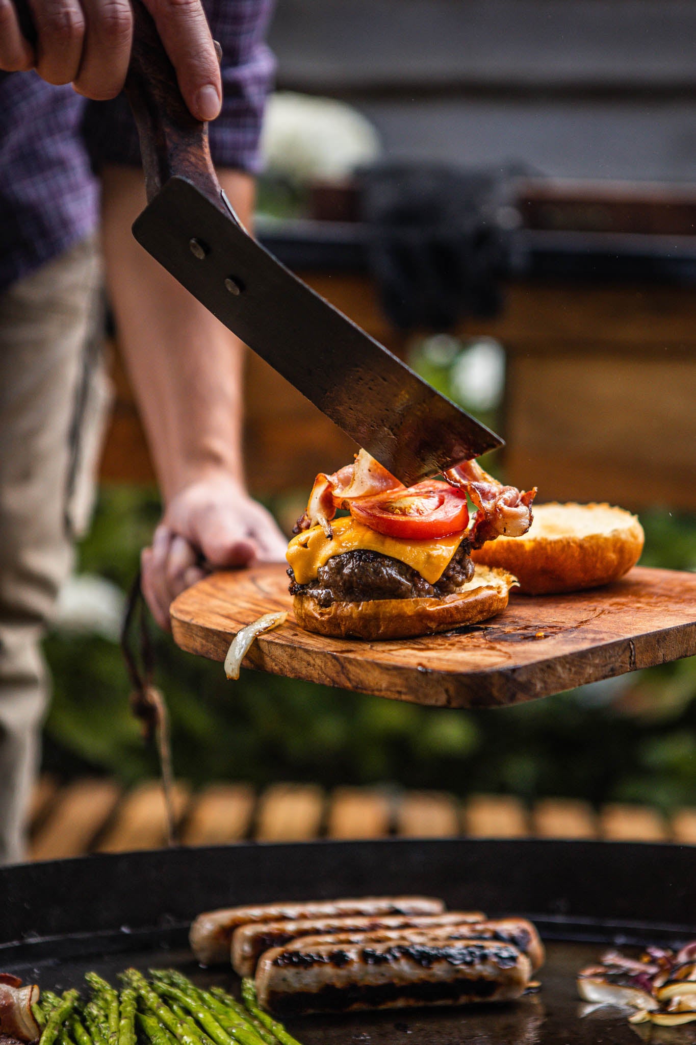 Close-up of a rustic BBQ spatula assembling a gourmet cheeseburger beside sizzling sausages on a flat-top outdoor barbecue grill