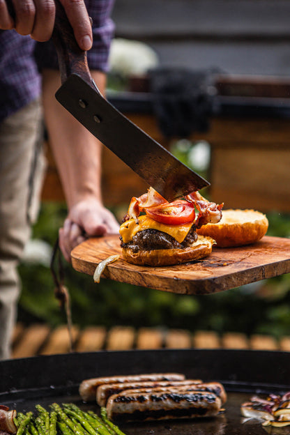 Close-up of a rustic BBQ spatula assembling a gourmet cheeseburger beside sizzling sausages on a flat-top outdoor barbecue grill