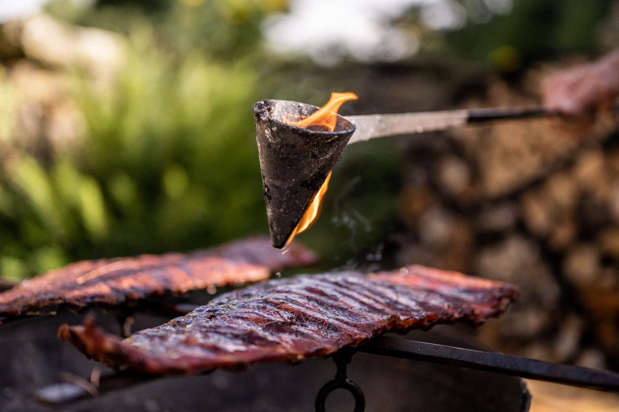 Close-up of a traditional flambadou suspended above smoked ribs with flames and smoke rising around it