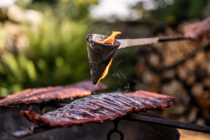 Close-up of a traditional flambadou suspended above smoked ribs with flames and smoke rising around it