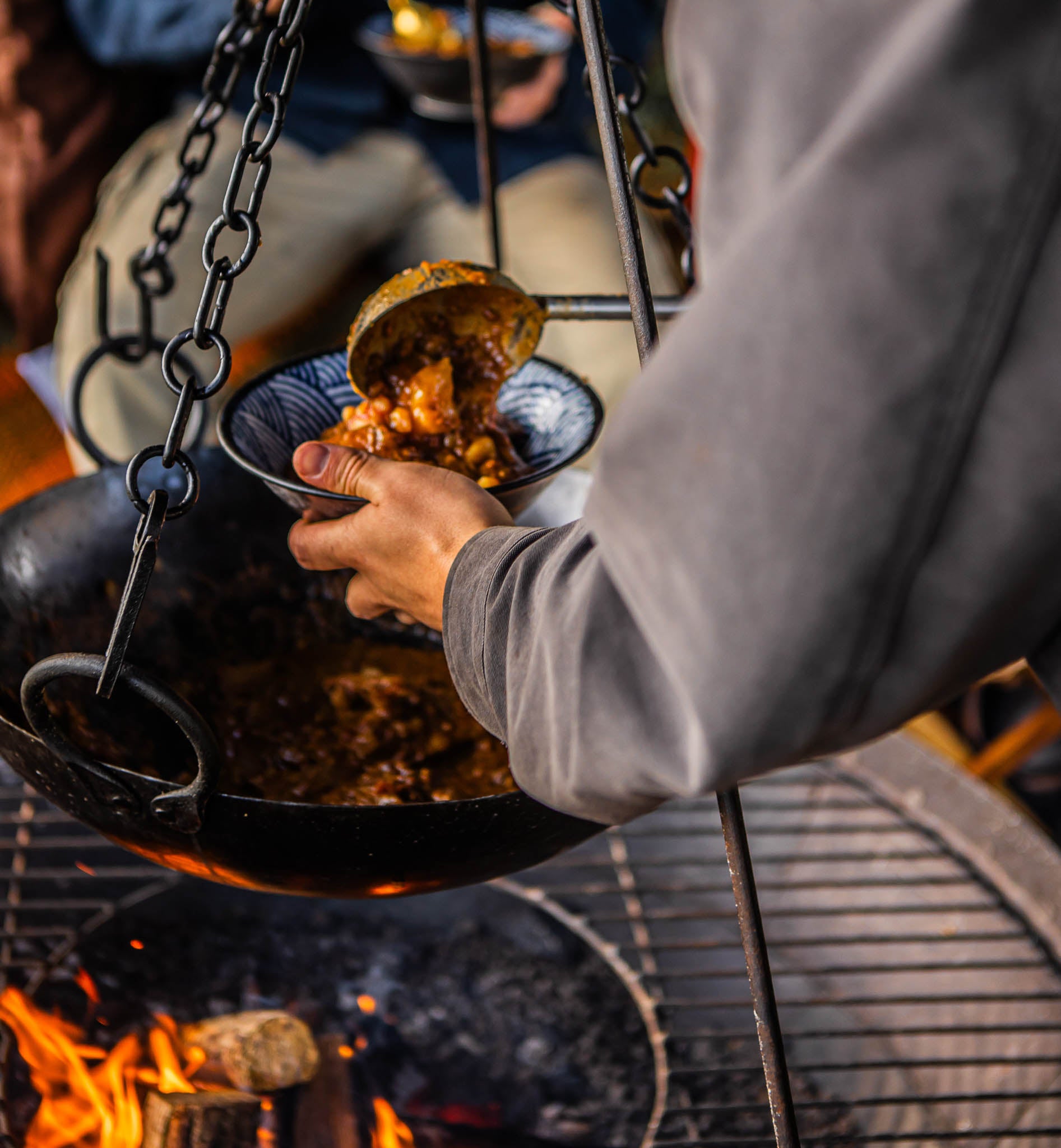 Long-handled stainless steel barbecue ladle serving slow-cooked outdoor food over an open fire pit