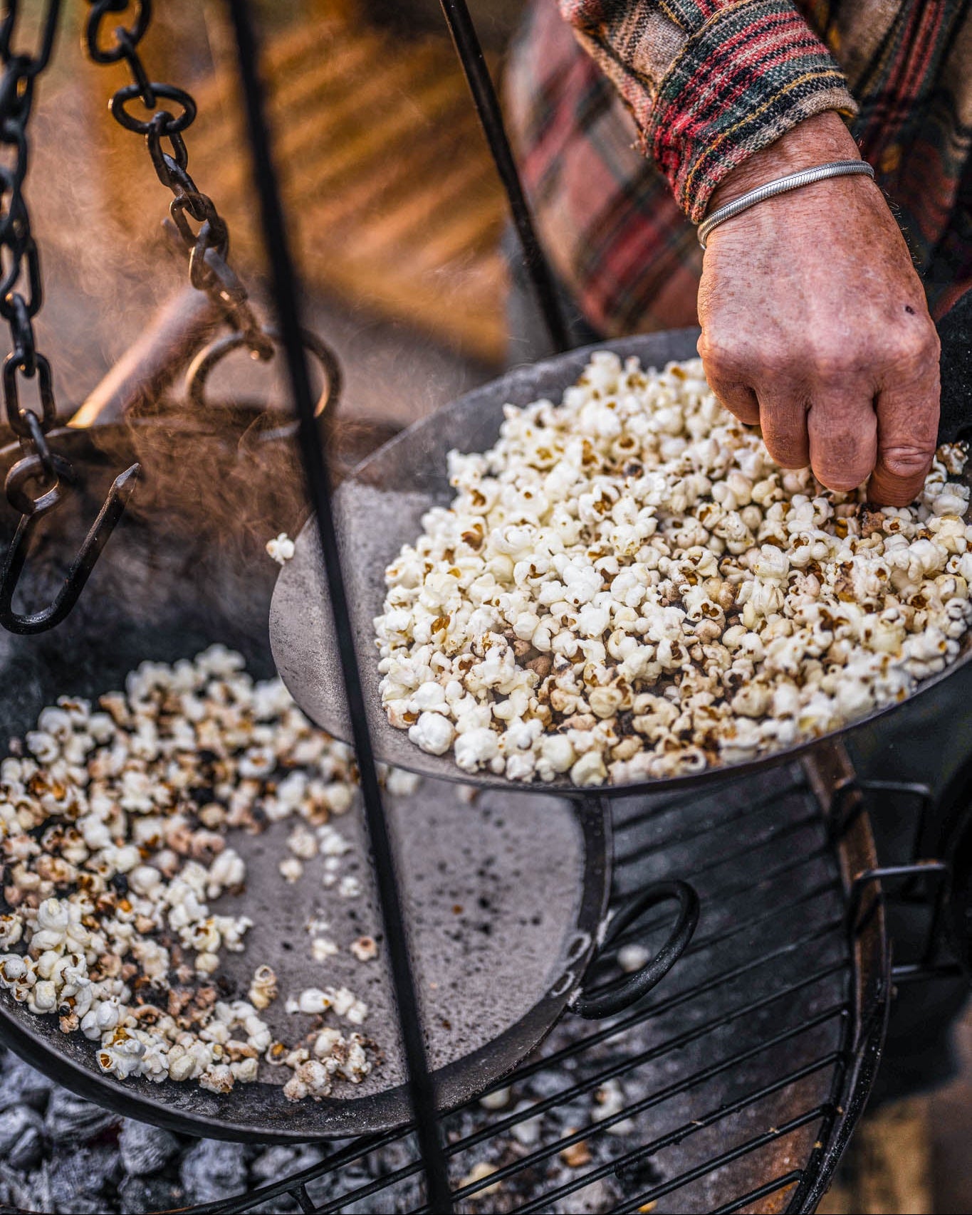Popcorn being cooked in a pan over an outdoor tripod roaster, with a hand stirring the kernels above hot coals