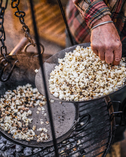 Popcorn being cooked in a pan over an outdoor tripod roaster, with a hand stirring the kernels above hot coals