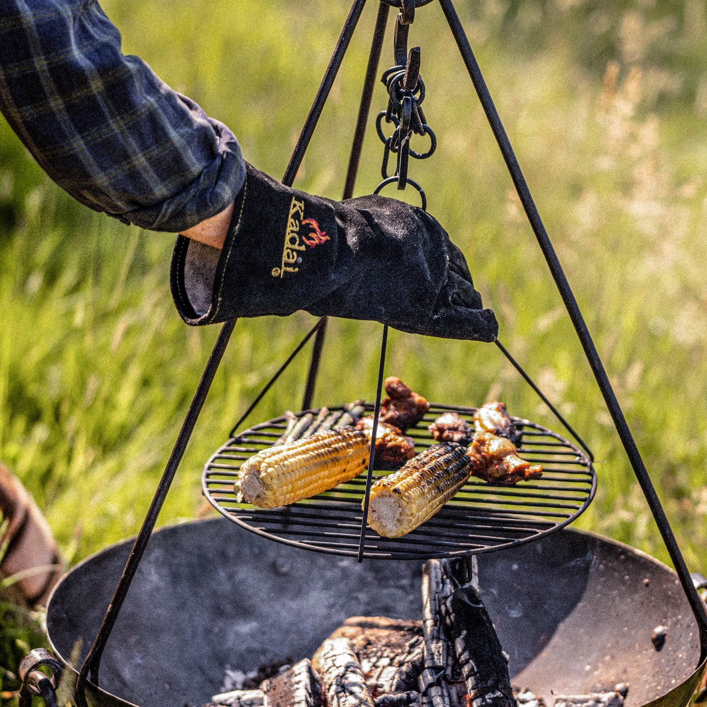 Person wearing heat-resistant kadai glove adjusting a hanging grill over an open fire