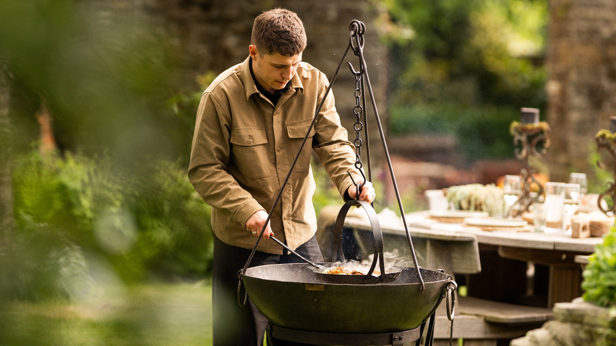 A person tends food cooking over a large black kadai fire bowl suspended from a metal tripod in a lush garden setting, using cooking tools while smoke rises from the pan