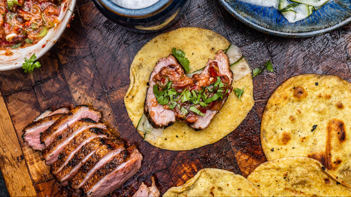 Overhead food scene on a rustic wooden board with a taco topped with sliced duck, salsa, and chopped green herbs, alongside sliced meat, tortillas, a bowl of salsa, and a blue plate of sliced cucumber.