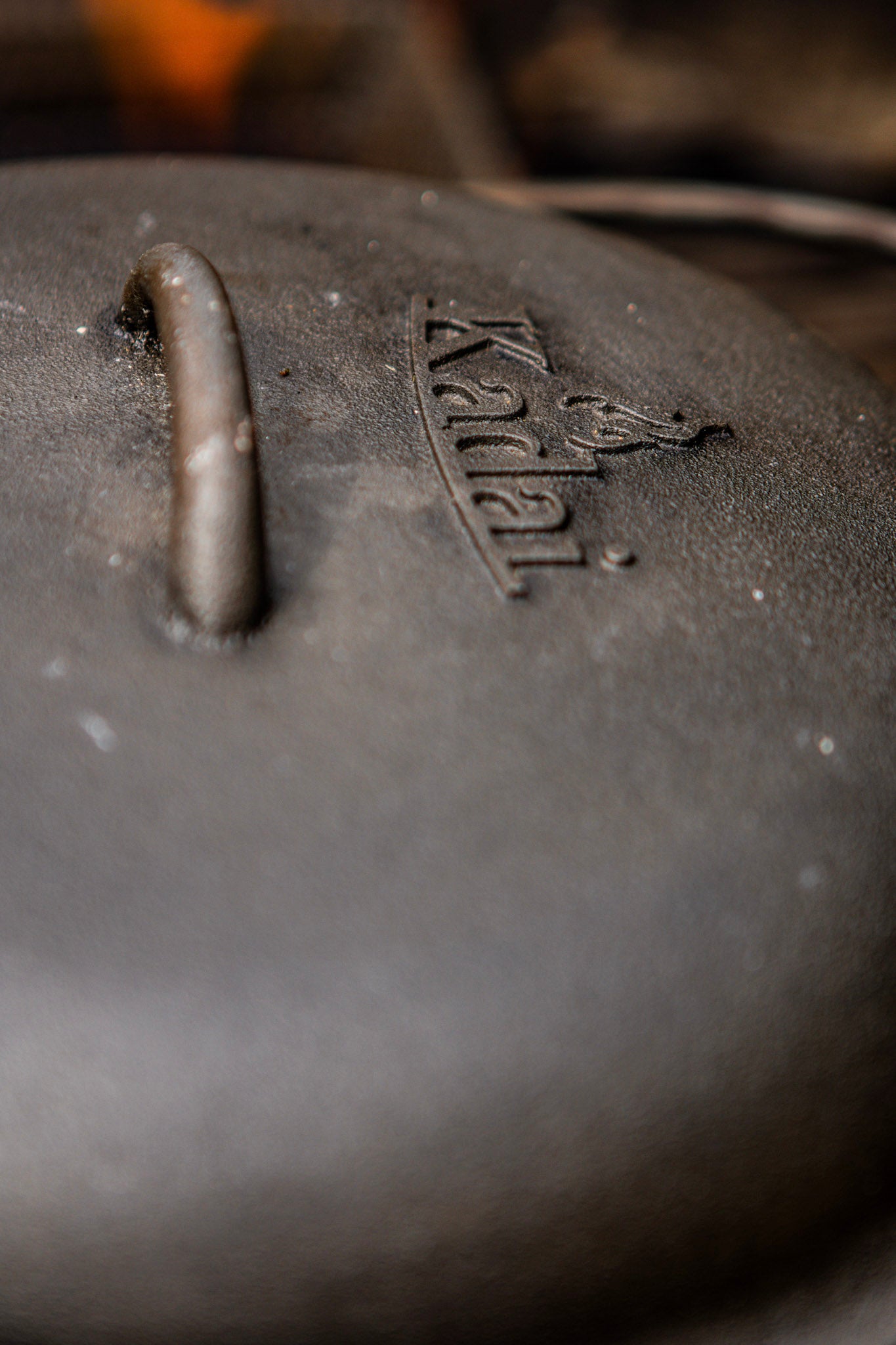 Close-up of a dark cast iron pot lid with a handle and the embossed brand name 'Kadai' on a blurred warm-toned background.