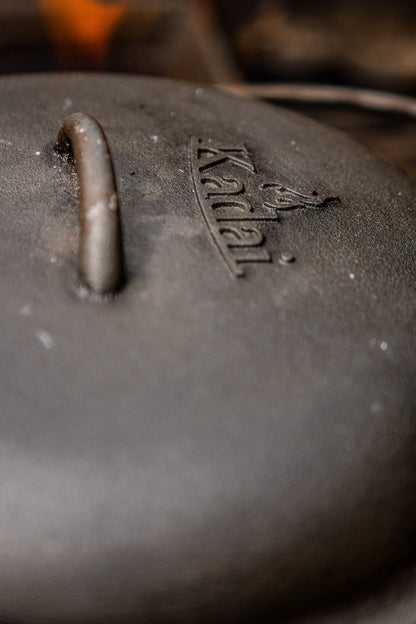Close-up of a dark cast iron pot lid with a handle and the embossed brand name 'Kadai' on a blurred warm-toned background.