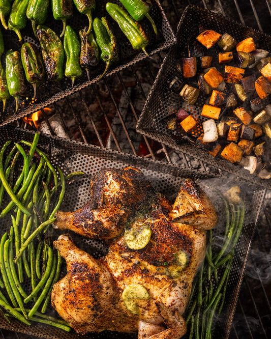 Assorted foods cooking on perforated BBQ grill trays over hot coals, including grilled chicken, green beans, peppers, and roasted vegetables with visible char and smoke.
