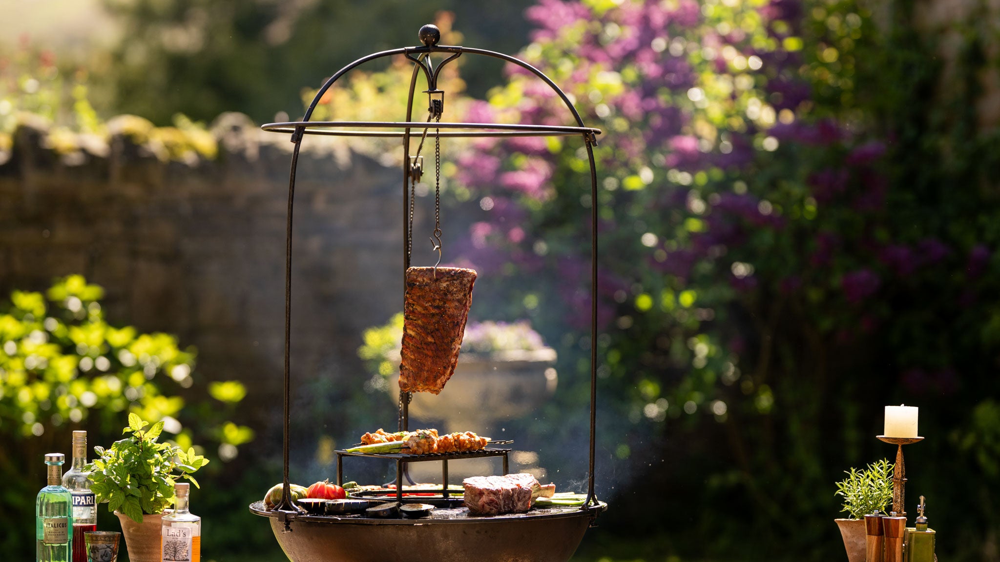 BBQ plancha grill with hanging live fire cooking system, featuring steaks, skewers and vegetables cooked over open flames in an outdoor garden setting