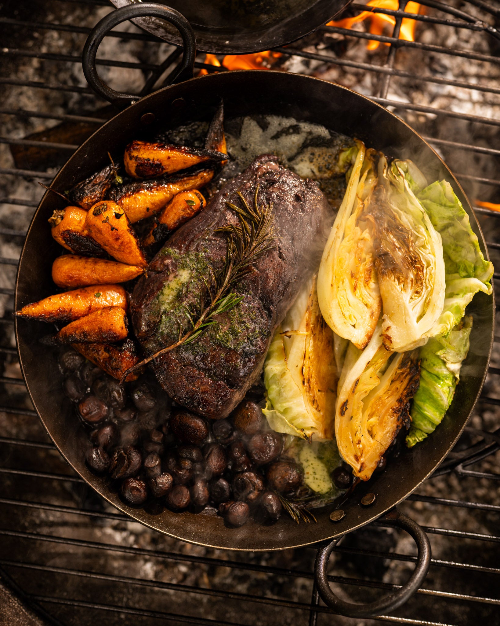BBQ skillet with grilled steak, charred cabbage, roasted carrots, and mushrooms cooking over an open fire grill.
