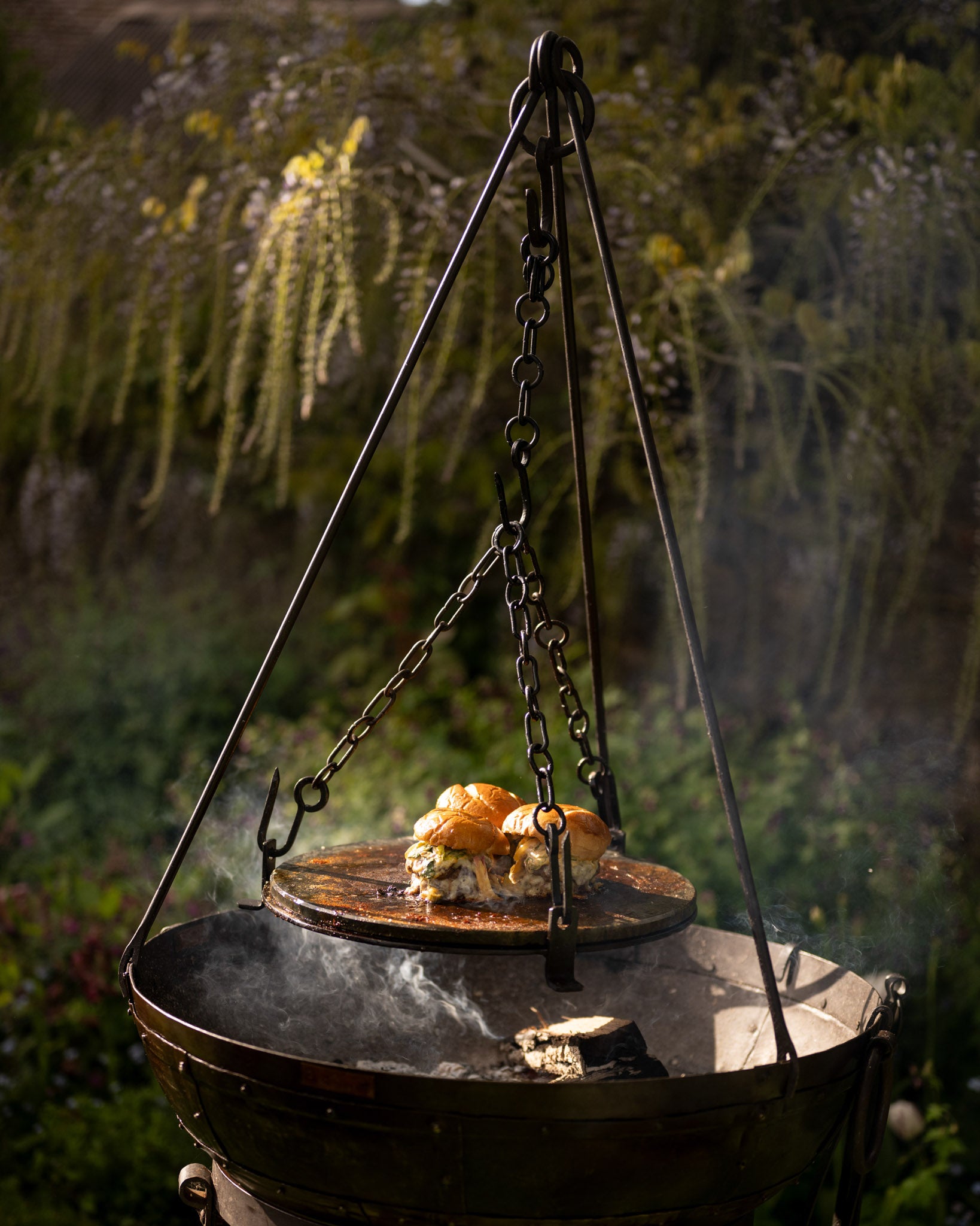 Stone griddle plate suspended over a Kadai firebowl barbecue with burgers cooking above hot coals in an outdoor garden setting