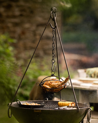 Outdoor tripod roaster with a whole chicken suspended above a fire pit alongside grilled corn and roasted vegetables.