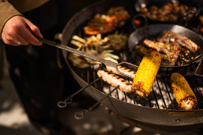 Hand using a metal grilling fork to roast corn over a large kadai cooking utensil with grilled vegetables and meat.