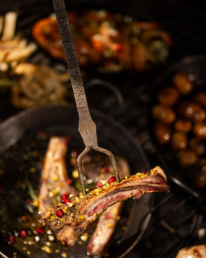 Grilled lamb chop held over a kadai cooking utensil with spices and herbs, surrounded by roasted dishes in a rustic kitchen setting.