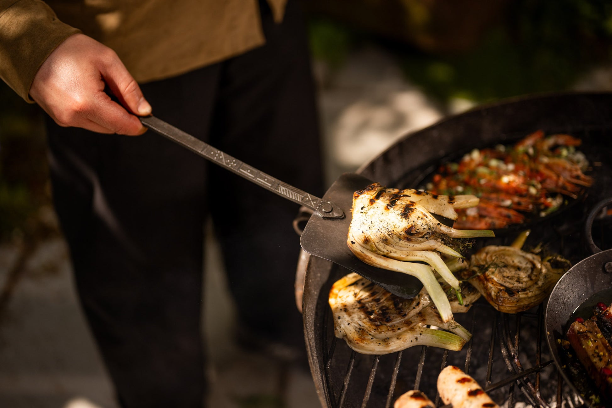 Person grilling charred Vegetable with a metal spatula over a large kadai cooking utensil filled with vegetables and roasted foods