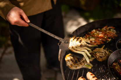 Person grilling charred Vegetable with a metal spatula over a large kadai cooking utensil filled with vegetables and roasted foods
