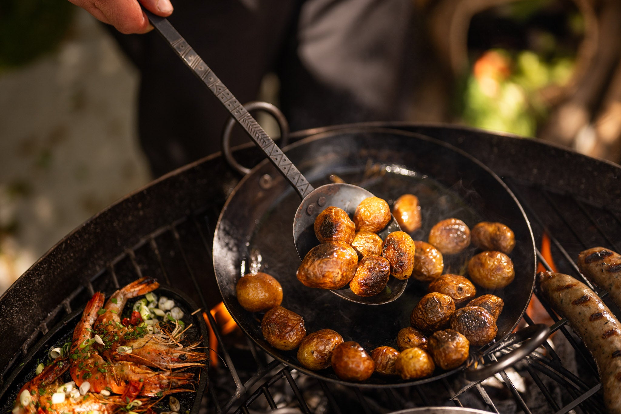 Kadai utensil scooping roasted potatoes from a pan with grilled prawns and sausages over an open flame.