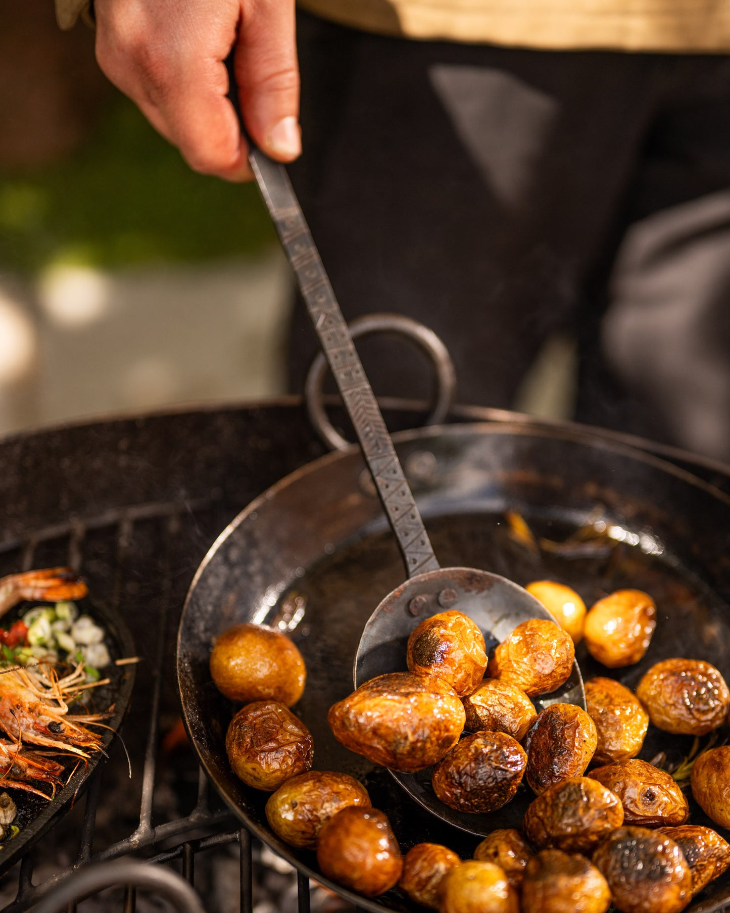 Hand-forged Kadai cooking utensil lifting roasted potatoes from a traditional fire bowl pan, designed for live fire cooking, outdoor barbecuing and durable open-fire food preparation