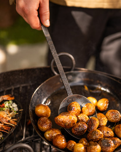 Hand-forged Kadai cooking utensil lifting roasted potatoes from a traditional fire bowl pan, designed for live fire cooking, outdoor barbecuing and durable open-fire food preparation