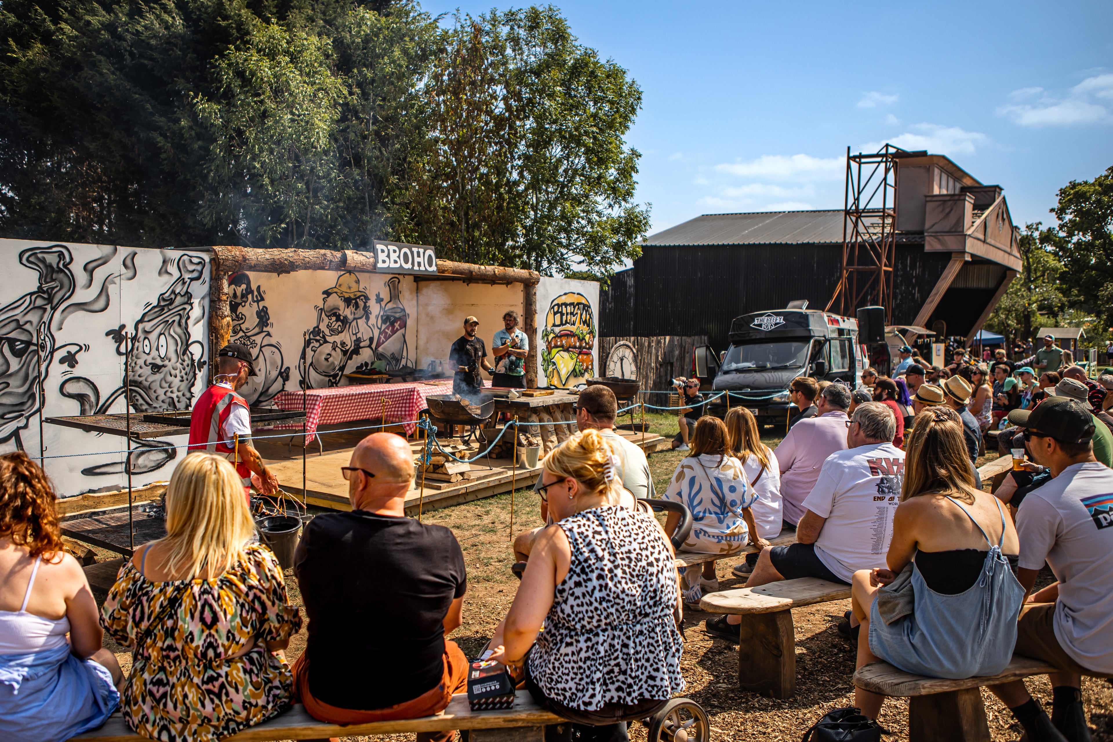 Outdoor event with people sitting on benches, a stage, and a truck with graffiti art.