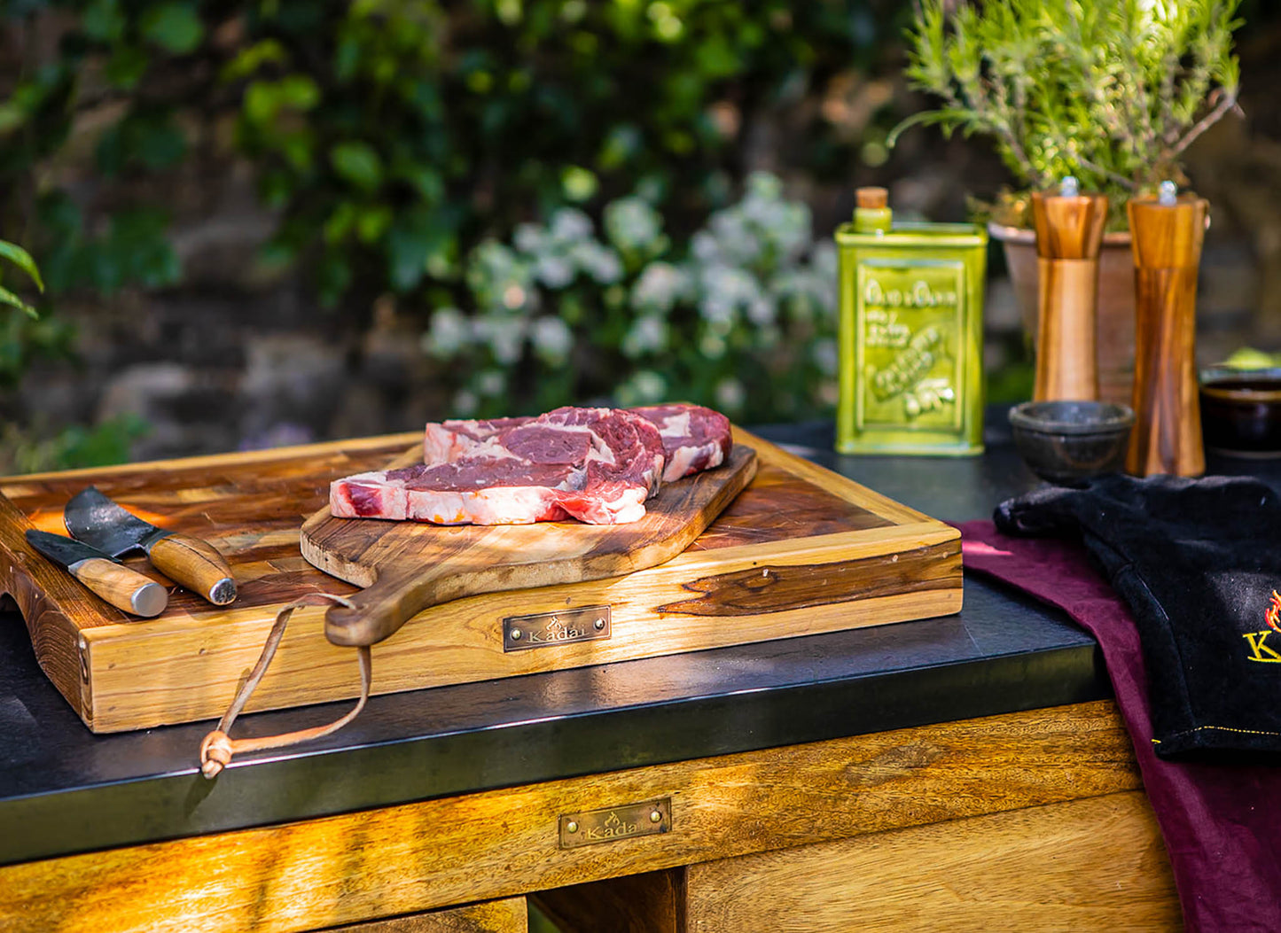 Large wooden butcher’s block with raw steaks on a rustic cutting board, set on an outdoor kitchen counter with herbs, oil tin, and grilling tools nearby.