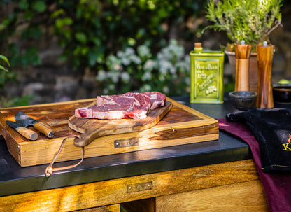 Large wooden butcher’s block with raw steaks on a rustic cutting board, set on an outdoor kitchen counter with herbs, oil tin, and grilling tools nearby.