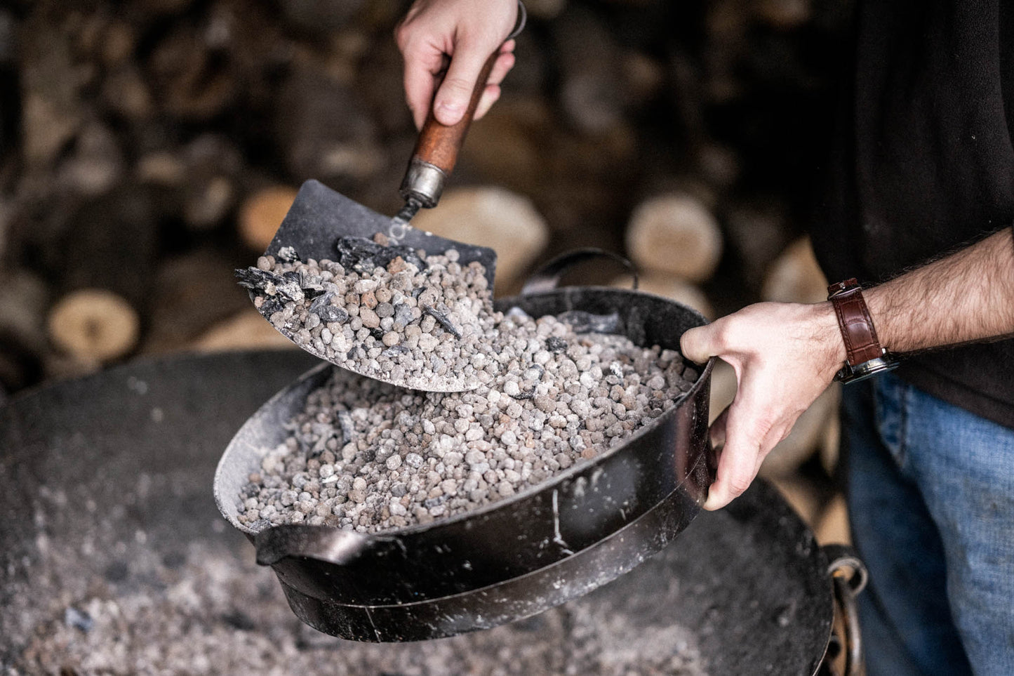 Person using a Kadai shovel with a wooden handle to transfer ash and embers into a black metal ash pan beside a fire bowl.