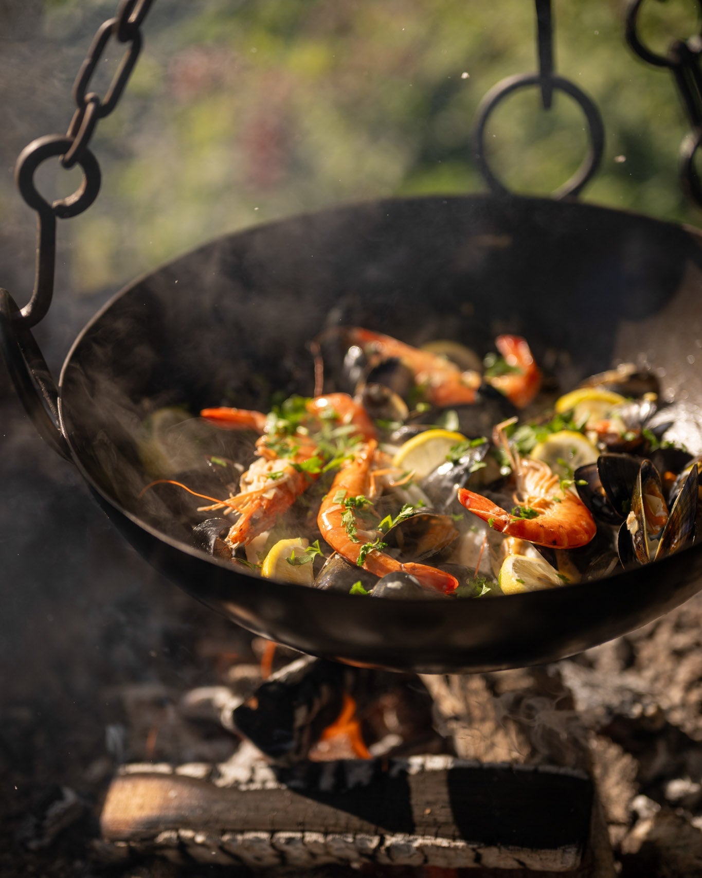 Seafood cooking in a hanging BBQ cooking bowl over an open fire, featuring prawns, mussels, lemon slices, and fresh herbs.