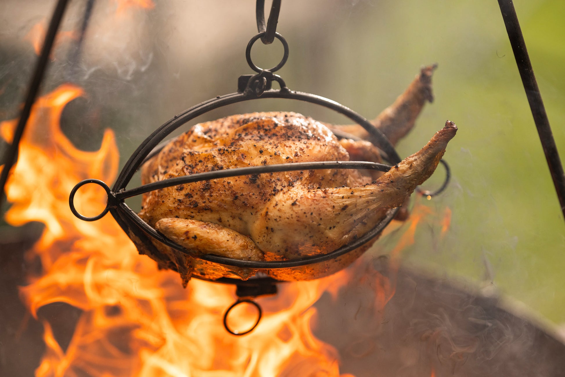 Whole chicken roasting in a hanging tripod roaster basket above open flames at an outdoor fire pit.