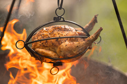 Whole chicken roasting in a hanging tripod roaster basket above open flames at an outdoor fire pit.